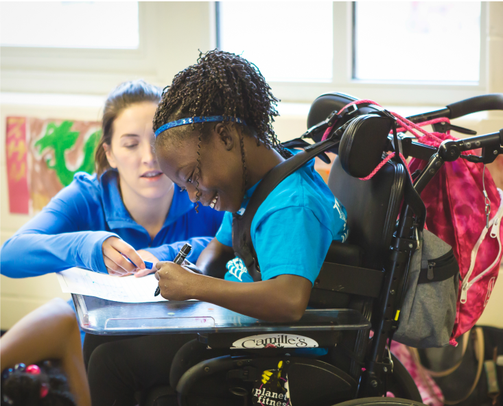 Smiling Girls on the Run participant in wheelchair writing as kneeling coach helps