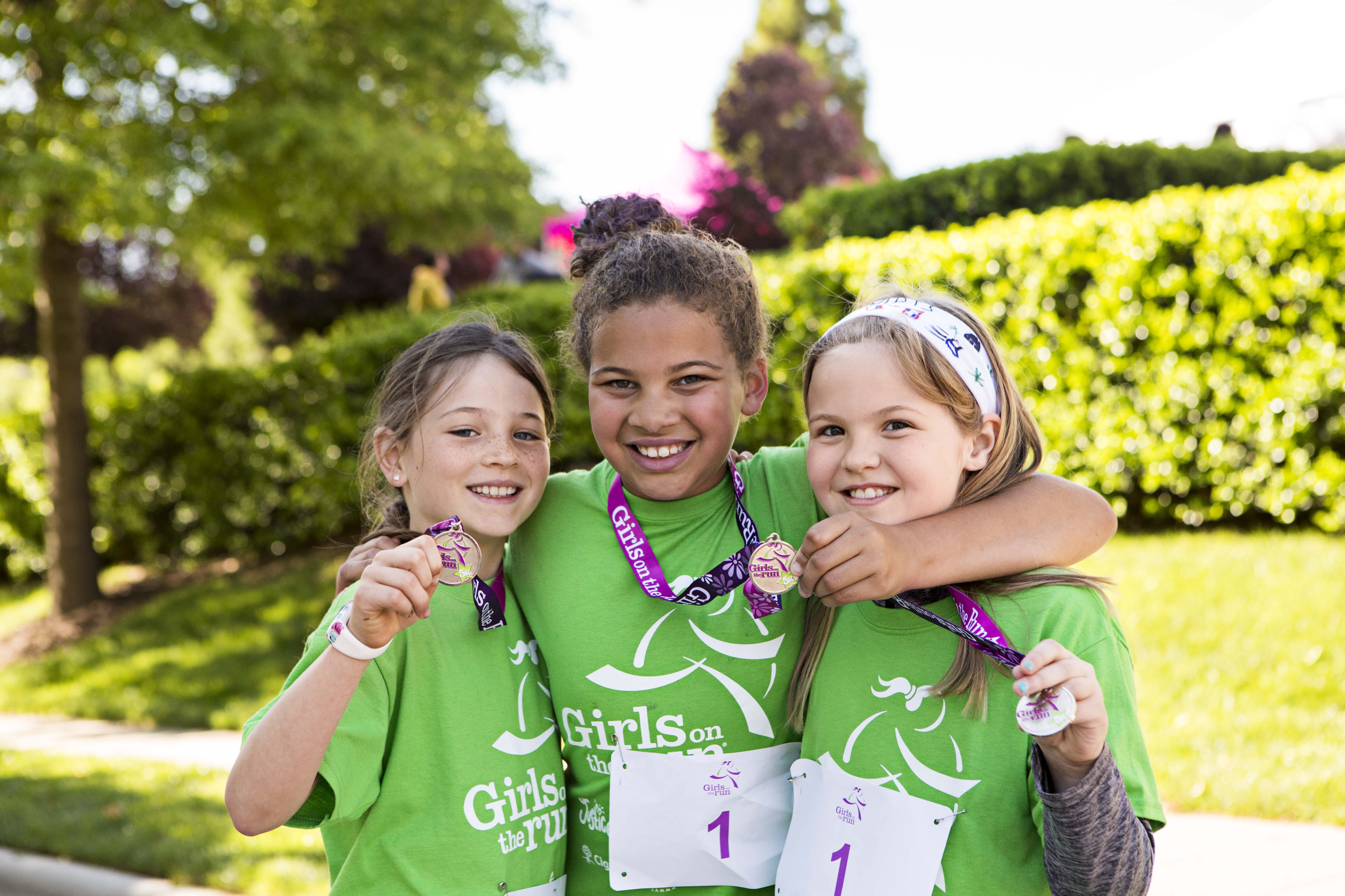 3 girls in green shirts smile while holding medals after the Girls on the Run 5K